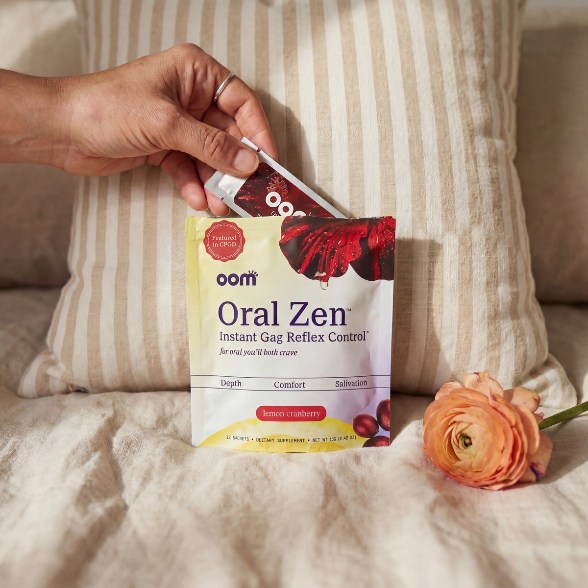 Hand pulling Oral Zen natural gag reflex supplement sachet from pouch against striped linen pillow in soft warm light