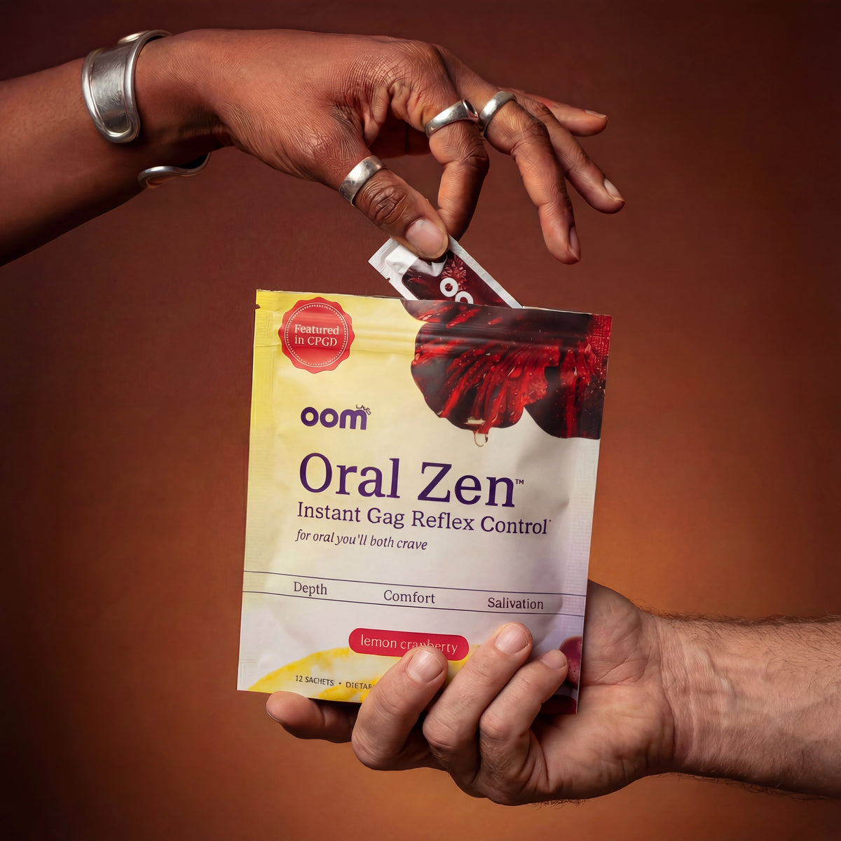 Hand pulling an Oral Zen instant gag reflex supplement sachet from the 12-pack pouch against a warm brown gradient. Woman's hand, black, with silver jewelry, and detailed skin, about 30 years old, pulls single sachet from the multi pouch. Multi pouch is held by white masculine hand from underneath, his hand entering the scene from the bottom right of the image.