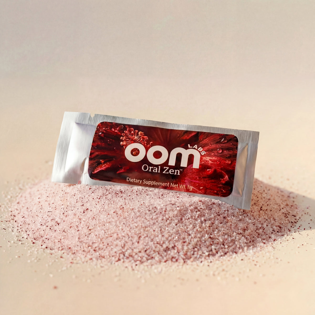 Oral Zen gag reflex control sachet resting in a pile of its fine pink lemon cranberry powder on a warm cream background. The powder is the fineness of table salt, light pink in color, with tiny dark maroon specks throughout. High intensity lighting from the front left creates a shimmer on the left side of the packet and a shadow on the right side. A shadow is cast on the back side of the 3-inch round pile of crystalline powder.
