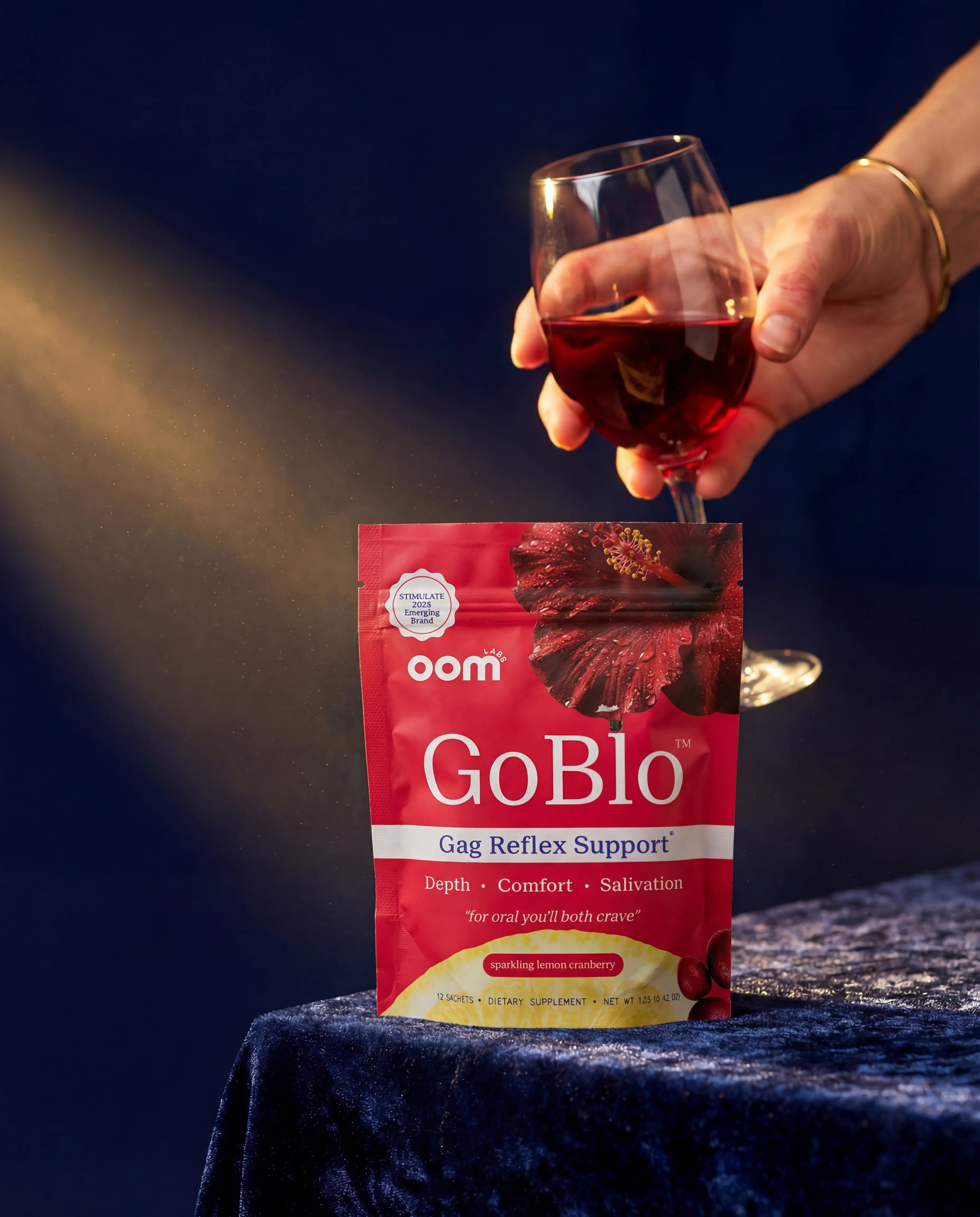 Hand holding a glass of red wine above a 'oom Goblo' package on a dark background