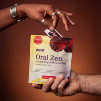 Hand pulling an Oral Zen instant gag reflex supplement sachet from the 12-pack pouch against a warm brown gradient. Woman's hand, black, with silver jewelry, and detailed skin, about 30 years old, pulls single sachet from the multi pouch. Multi pouch is held by white masculine hand from underneath, his hand entering the scene from the bottom right of the image.