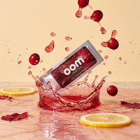 Oral Zen lemon cranberry gag reflex supplement sachet splashing into cranberry water with lemon slices and hibiscus. Five cranberries are caught mid-air as they fall into a light pink cranberry juice pool. Two wheels of lemon sit at water-level in the pink pool. The Oral Zen sachet's front label faces the viewer directly. A few hibiscus petals float on the liquid.