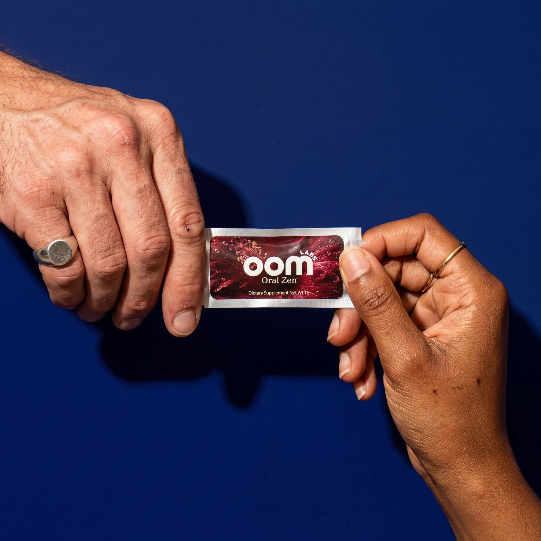 Two hands exchanging an Oral Zen instant gag reflex control sachet against a deep navy background. The left hand is male, 50 years old, with a well-worn silver ring on his pinky finger. He has slightly worked hands. The female's hand enters from the bottom right. Her skin is mid-tone black, with a few black freckles. She has well-manicured short unpainted nails, and a thin gold ring on her pointer finger. A dark shadow is cast on the navy background from the overhead lighting.