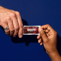 Two hands exchanging an Oral Zen instant gag reflex control sachet against a deep navy background. The left hand is male, 50 years old, with a well-worn silver ring on his pinky finger. He has slightly worked hands. The female's hand enters from the bottom right. Her skin is mid-tone black, with a few black freckles. She has well-manicured short unpainted nails, and a thin gold ring on her pointer finger. A dark shadow is cast on the navy background from the overhead lighting.