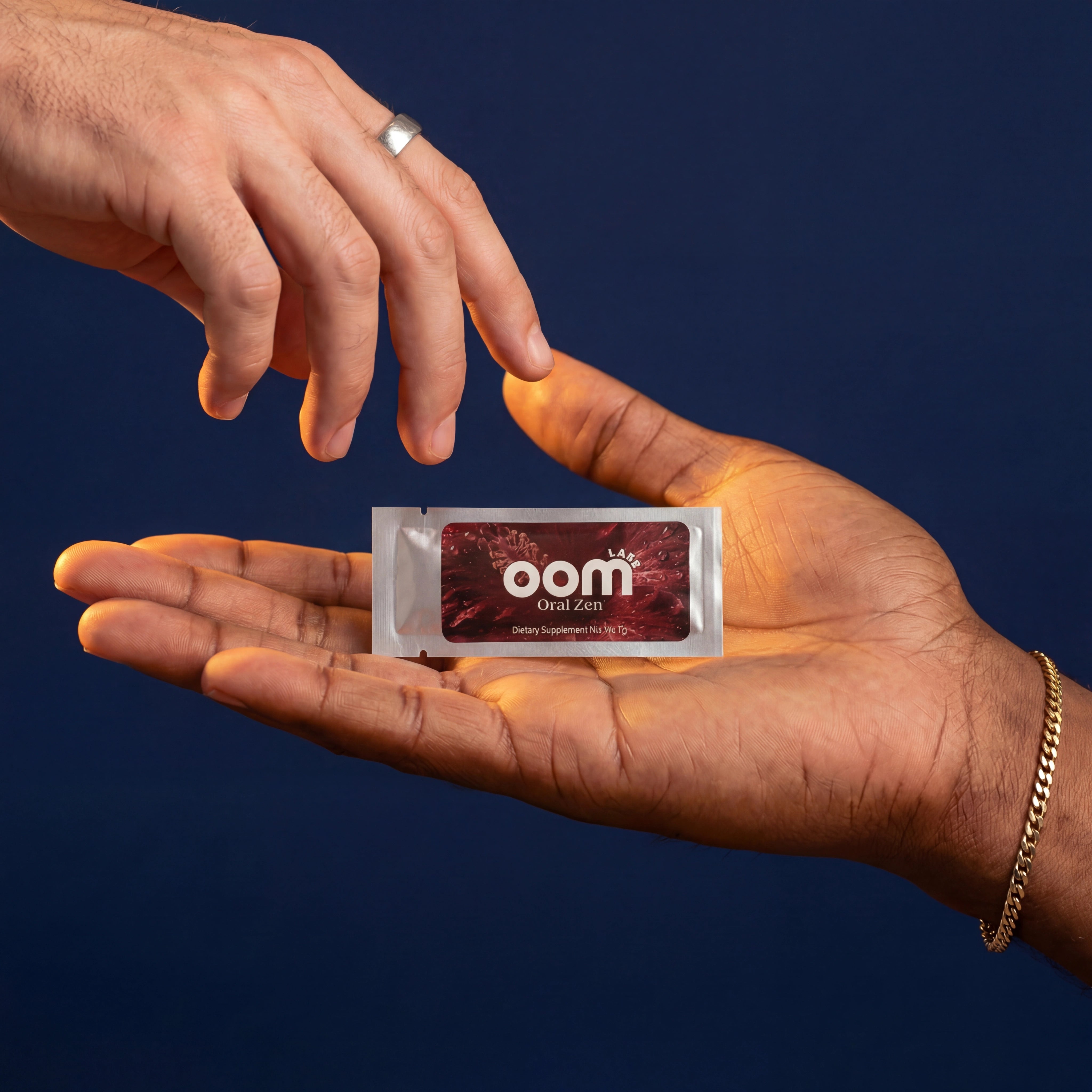 Oral Zen all-natural gag reflex supplement sachet resting on an open palm with another hand reaching for it on navy background. The hand holding the sachet comes from the bottom right of the screen. It is a large male hand, dark skin, with a fine gold chain. The other hand comes from the upper left. It is a smaller male hand, right hand, mid-density hair on the back of his palm. He wears a dulled silver ring on his pointer finger.
