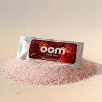 Oral Zen gag reflex control sachet resting in a pile of its fine pink lemon cranberry powder on a warm cream background. The powder is the fineness of table salt, light pink in color, with tiny dark maroon specks throughout. High intensity lighting from the front left creates a shimmer on the left side of the packet and a shadow on the right side. A shadow is cast on the back side of the 3-inch round pile of crystalline powder.