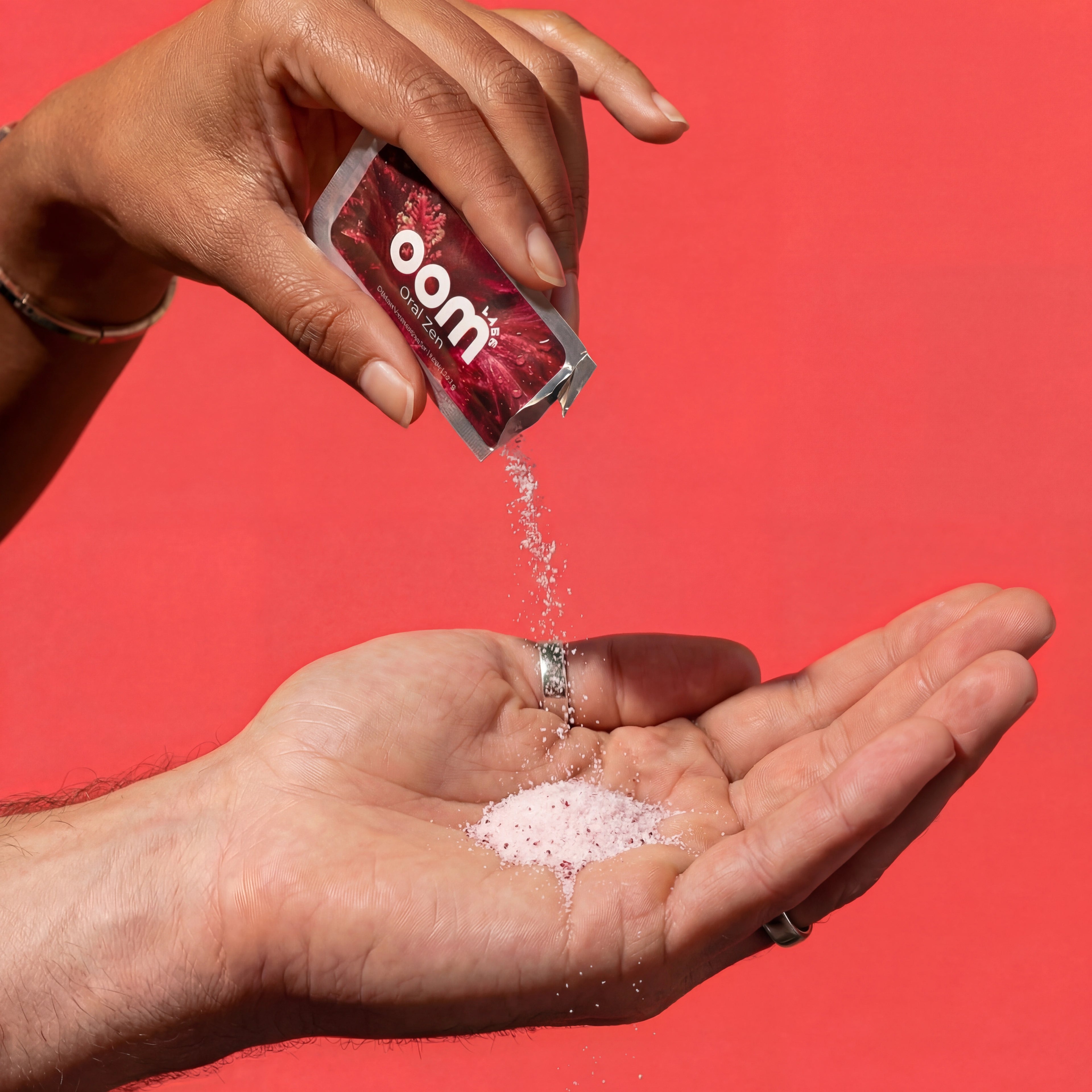 Oral Zen lemon cranberry gag reflex powder being poured from a sachet into a cupped hand against a coral background. The hand pouring the crystalline powder from the small sachet is a smooth-skinned black woman with well-groomed short nails and a gold bangle. The hand receiving the powder is a 35 year old white male with silver rings on his thumb and ring finger. Both hands come from the left side of the image.
